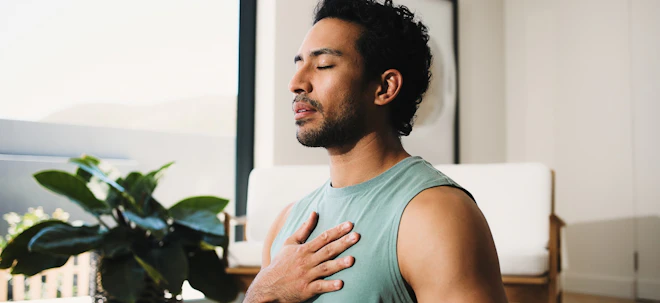 A man sits with his eyes closed and one hand resting on his chest, breathing slowly in a sunlit room