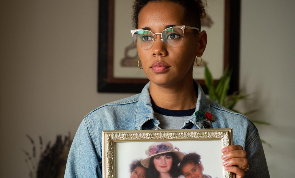 African-American woman with denim jacket and wearing glasses. Looking off to the right of the camera. Holding a photo of her mother and Alica's siblings when they were children.
