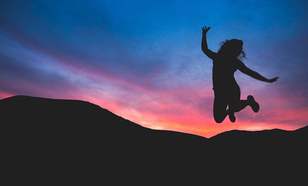 Silhouette of woman jumping in the air. Background is a silhouette of mountains wit a blue and pink sunset sky.