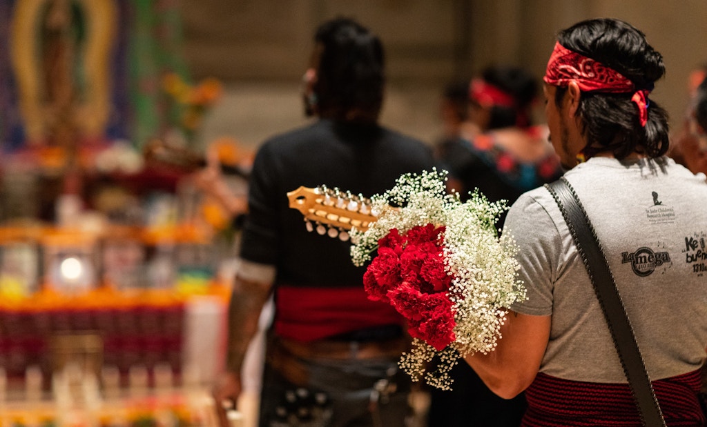 Man wearing red bandana and holding red rose bouquet. Background is a Dia de los Muertos altar.