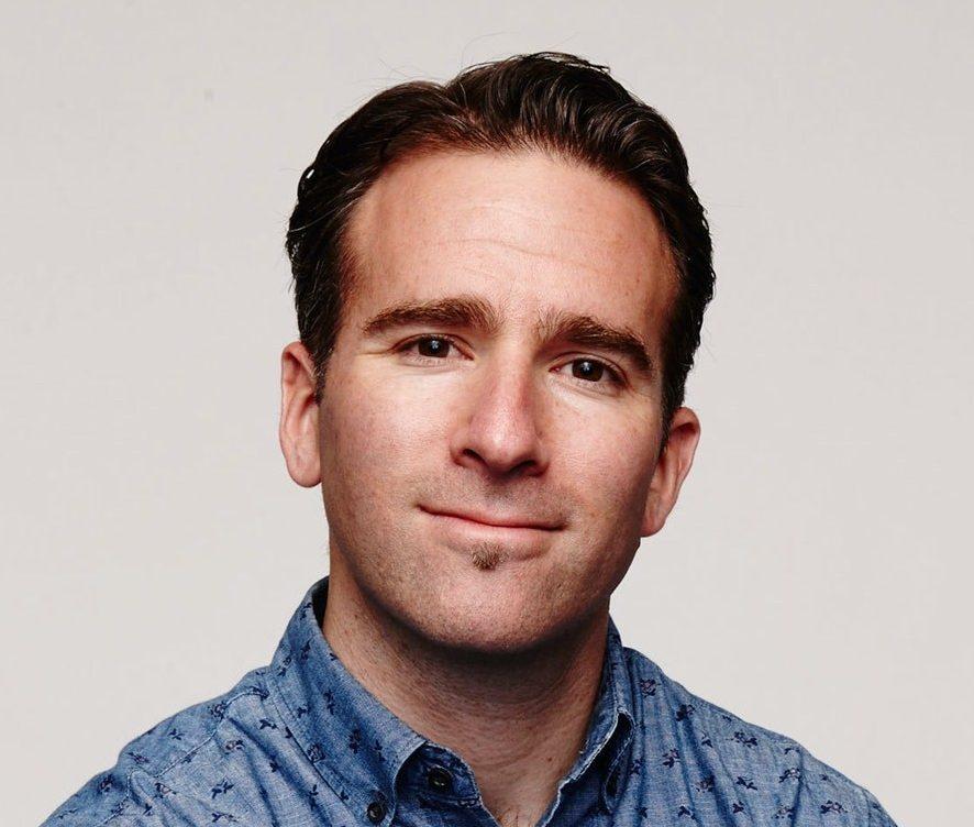 White man with blue button-down shirt looking at the camera. Sitting casually on metal chair.
